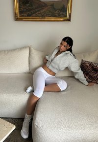 Sweat à capuche gris court, leggings blancs et bottes de couleur claire. Canapé beige clair texturé avec un design confortable. Coussin à imprimé léopard à proximité.