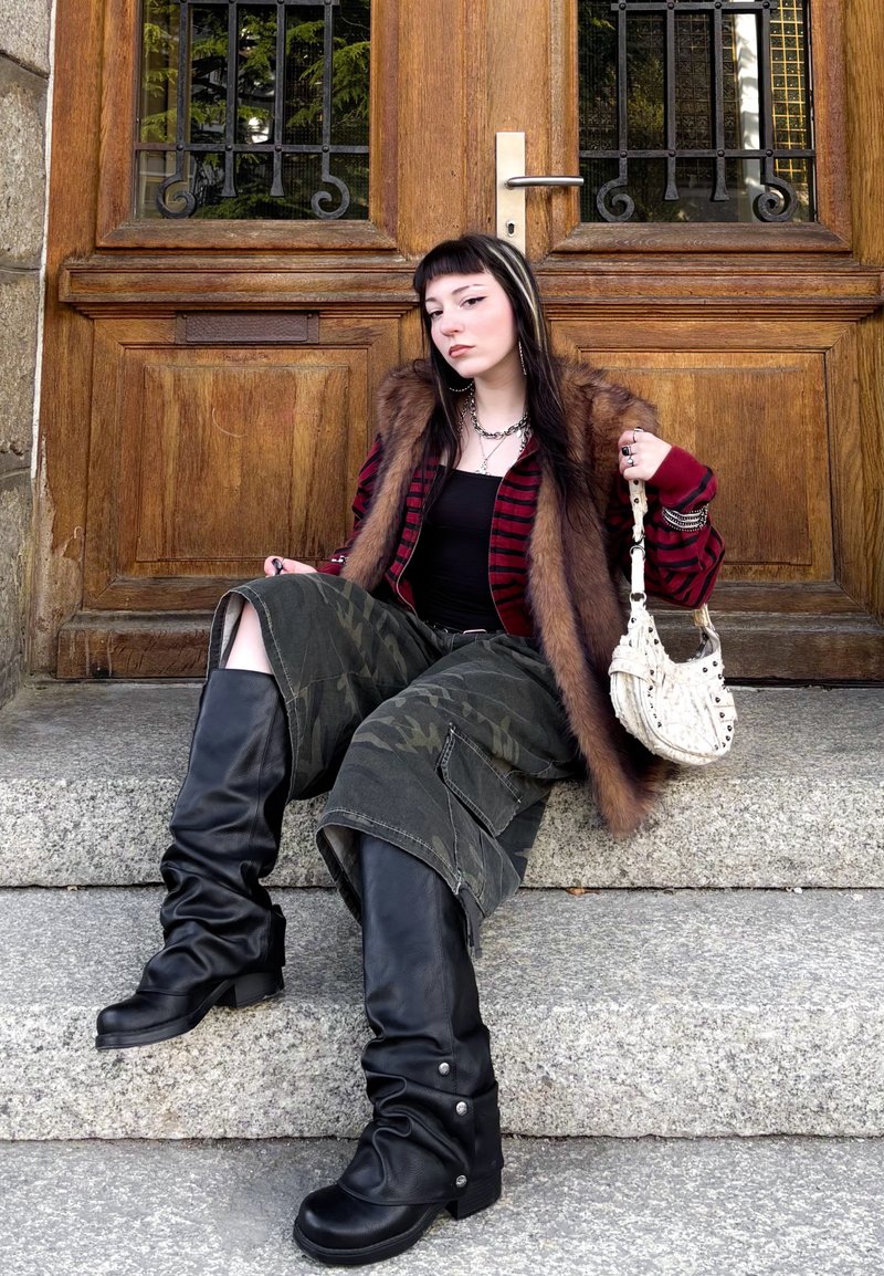 Jeune femme aux cheveux noirs et blancs assise sur des marches en pierre devant une porte en bois, portant un gilet en fourrure, un pantalon camouflage, des bottes noires, et tenant un sac à main blanc.
