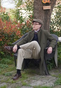 Zelený vlnený kabát, námornícky sveter, béžové nohavice, hnedé topánky. Osoba sedí na čiernej stoličke v záhrade medzi rastlinami a stromom.