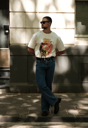 Camiseta blanca de gran tamaño con un estampado gráfico, jeans azules y zapatos de cuero negros. El hombre lleva gafas de sol negras y está de pie de manera casual.