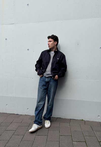 Chaqueta navy sobre una camisa a rayas, combinada con jeans azules y zapatillas blancas. El fondo es un concreto liviano gris claro.