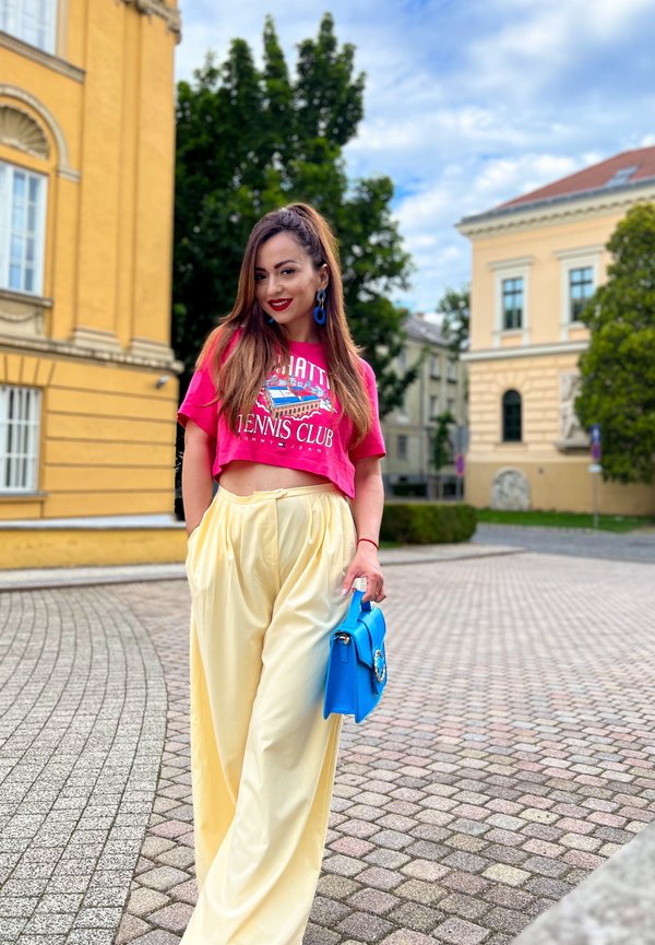 T-shirt cropped rosa com design gráfico, calças largas amarelas e bolsa azul. O cenário apresenta pavimento de paralelepípedos e edifícios.