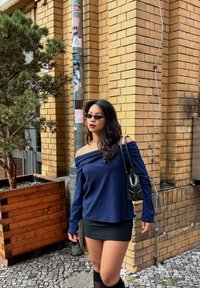Top off-shoulder blu navy, minigonna nera, calze al ginocchio e una borsa nera. Posizionata contro un muro di mattoni gialli con della vegetazione.