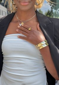 Λευκό strapless τοπ, μαύρη σακάκι, χρυσό βραχιόλι και στρωτές χρυσές αλυσίδες με καρδιά και διακοσμητικό κρεμαστό. Λείες υφές και λαμπερές φινιρίσματα.