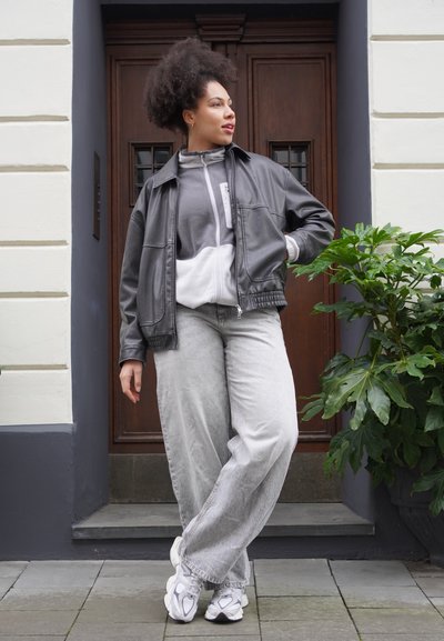 Cazadora bomber de cuero negro, chaqueta de forro polar gris con cremallera, jeans de pierna ancha grises y zapatillas blancas. De pie contra una puerta de madera con plantas.
