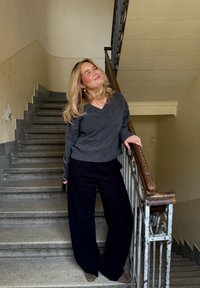 Grey knitted jumper with a V-neck, teamed with black wide-leg trousers. Standing on stairs with a wooden handrail in a light-coloured hallway.
