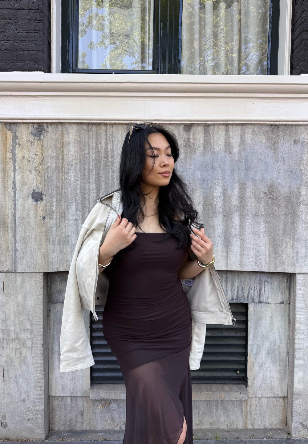 Abito nero attillato con una texture arricciata, abbinato a una giacca di pelle grigia chiara. Capelli intrecciati e una finestra con dettagli neri sullo sfondo.