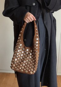 Bolsa de couro castanho com um topo curvado, adornada com tachas prateadas de vários tamanhos, com uma única alça de ombro.