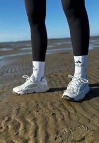 Chaussures de sport blanches avec des accents turquoise, en maille texturée et avec des lacets. Leggings noirs et chaussettes blanches avec des rayures noires sur une surface sableuse.