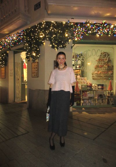 Top de punto de manga corta rosa claro combinado con una falda midi gris. Guantes y tacones negros. Cartera de mano plateada brillante. Fondo de escaparate festivo.