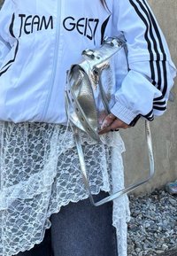 Biała kurtka typu zip z czarnymi paskami i napisem "TEAM GEIST", nałożona na białą koronkową sukienkę i dżinsy. Metaliczna srebrna torba na ramię.