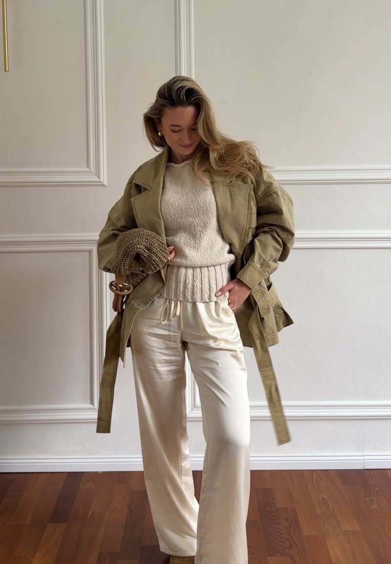 Femme aux longs cheveux portant un pull en maille beige, un pantalon large en satin blanc et une veste oversize olive, tenant un sac à main tressé marron.
