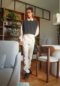 Grå tygblus med nitar i kanten över en vit skjorta med krage, kombinerat med krämfärgade byxor och svarta skor, placerad i en modern interiör.