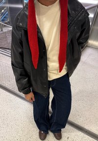 Veste en cuir noire avec une écharpe rouge en tricot, pull crème, et jean bleu à jambes larges. Des chaussures marron complètent la tenue.