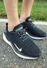 Os sapatos atléticos pretos apresentam um cabedal em malha com perfurações, atacadores cinzas e um logótipo swoosh branco, repousando sobre uma superfície cinza texturizada.