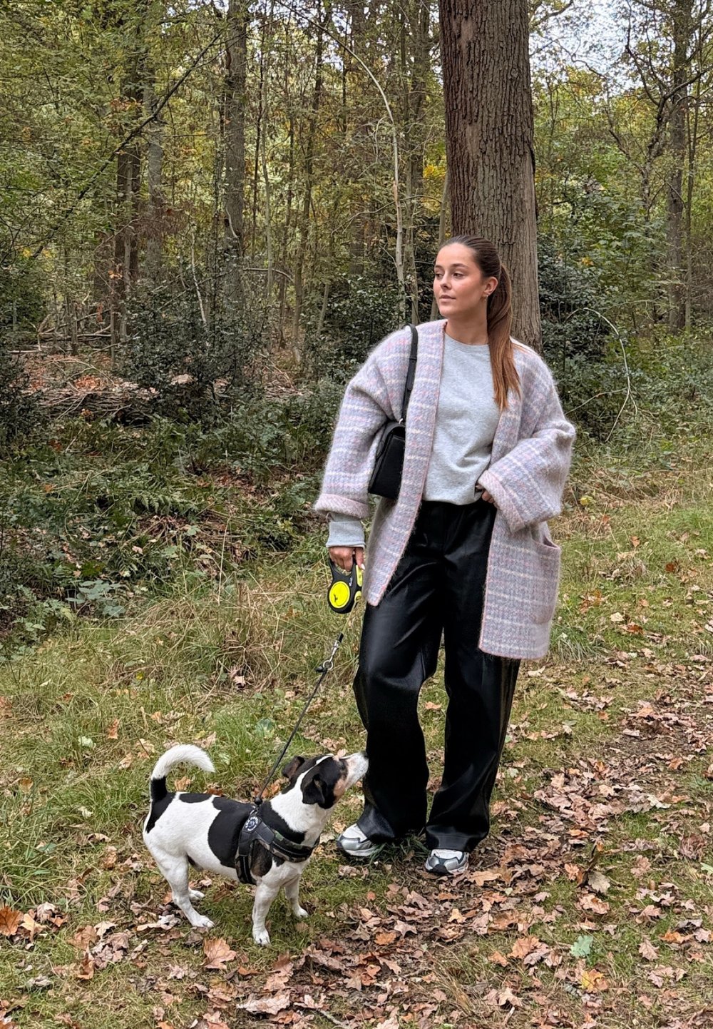 Un long cardigan texturé de couleur rose clair porté sur un sweat-shirt gris, assorti à un pantalon large noir. Un petit chien en laisse se tient à proximité.
