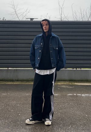 Joven con sudadera negra con capucha, chaqueta de mezclilla, pantalones deportivos negros de Adidas y zapatillas, de pie sobre pavimento mojado frente a una cerca gris.