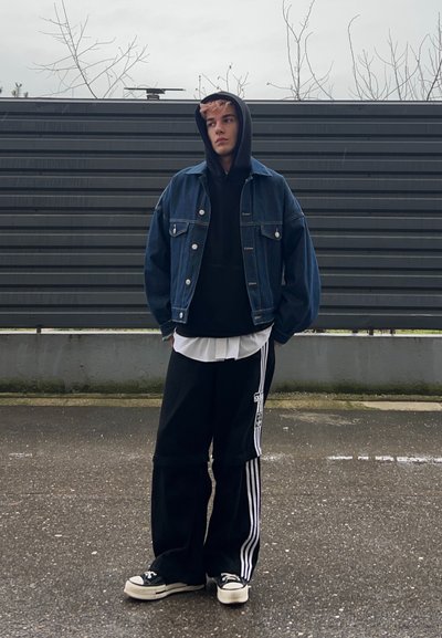 Joven con sudadera negra con capucha, chaqueta de mezclilla, pantalones deportivos negros de Adidas y zapatillas, de pie sobre pavimento mojado frente a una cerca gris.