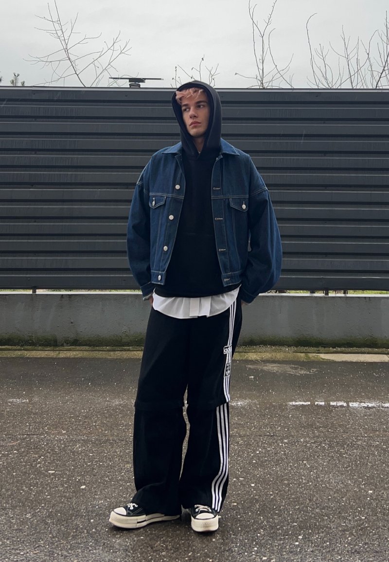 Jeune homme portant un sweat à capuche noir, une veste en jean, un pantalon de survêtement Adidas noir et des baskets, debout sur un trottoir mouillé devant une clôture grise.