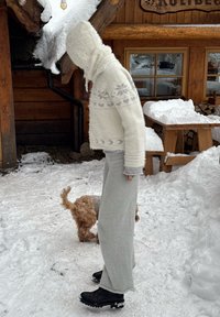 Fehér gyapjú pulóver kapucnival és hópehely mintával, világosszürke nadrággal és fekete csizmával, havas kültéri környezetben.