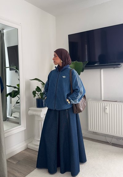 Chaqueta de denim con tres rayas blancas, logo bordado en el pecho, combinada con una falda de denim larga. Bolsa marrón y plantas verdes de fondo.