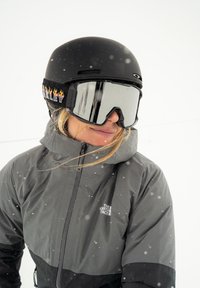 Zwarte skihelm met vlammen op het bandje, gespiegelde skibril en grijze jas met een zwart onderste deel met een logo. Sneeuw valt.