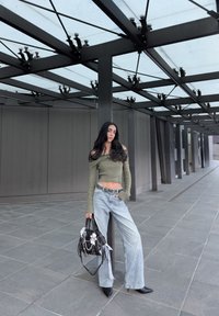 Haut en tricot vert olive à épaules dénudées, jean ample taille haute bleu clair, sac noir avec embellissements, escarpins noirs à bout pointu, arrière-plan architectural moderne.