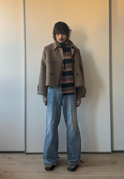 Persona con cabello oscuro que lleva una chaqueta marrón a cuadros, bufanda a rayas, vaqueros azules de pierna ancha y zapatos negros puntiagudos, de pie en el interior junto a una pared.