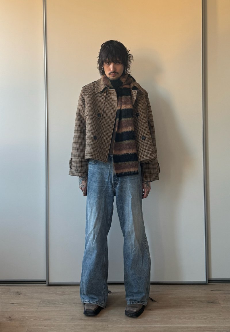 Tumejuukseline inimene kannab pruuni ruudulist jopet, triibulist salli, laiade säärtega siniseid teksapükse ja musti teravateotsalisi kingi, seistes siseruumis seina ääres.