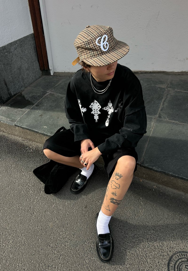 Jeune personne portant une casquette à motifs, un t-shirt à manches longues noir, un short noir, des chaussettes blanches, des chaussures noires, assise sur le trottoir montrant des tatouages sur la jambe gauche.