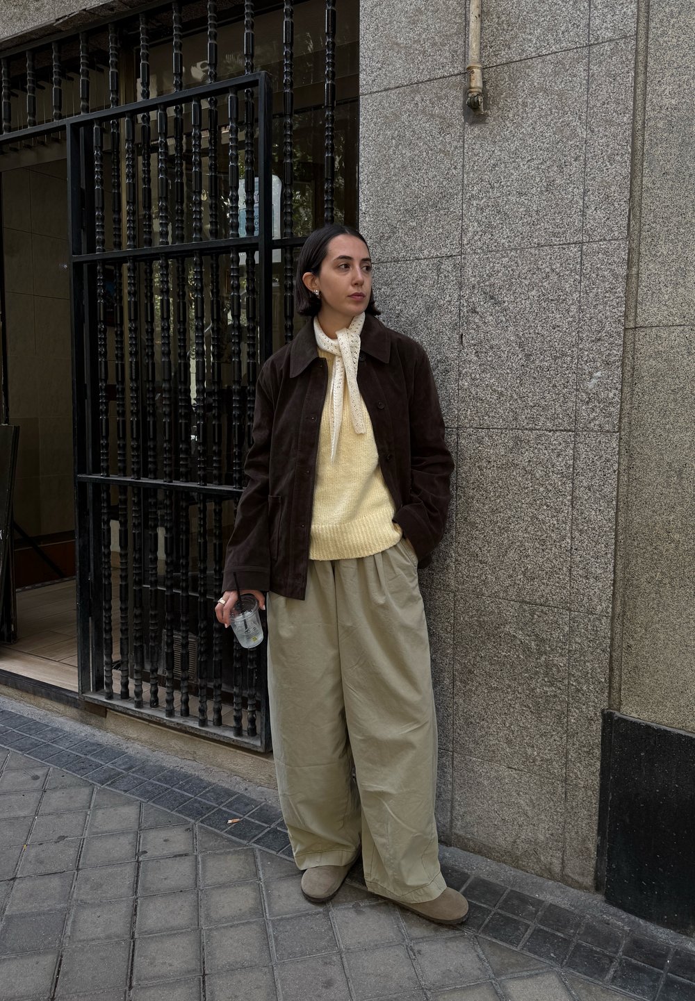 Brown suede jacket, light yellow knitted sweater, loose beige trousers, grey suede shoes, holding a transparent cup, standing against a textured wall.