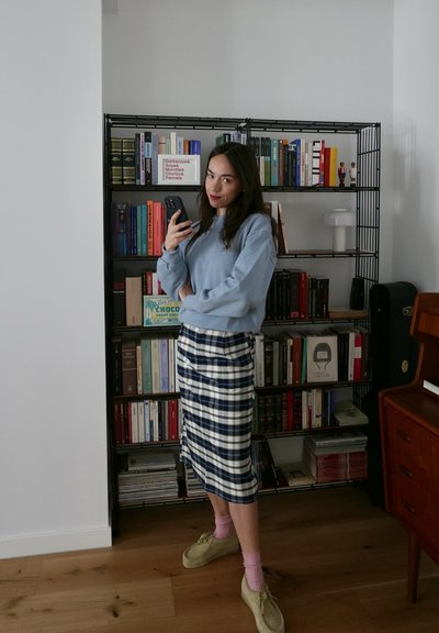 Sudadera azul clara, falda midi a cuadros en blanco y azul oscuro, calcetas rosas y zapatos beige. Estantería al fondo con libros variados.