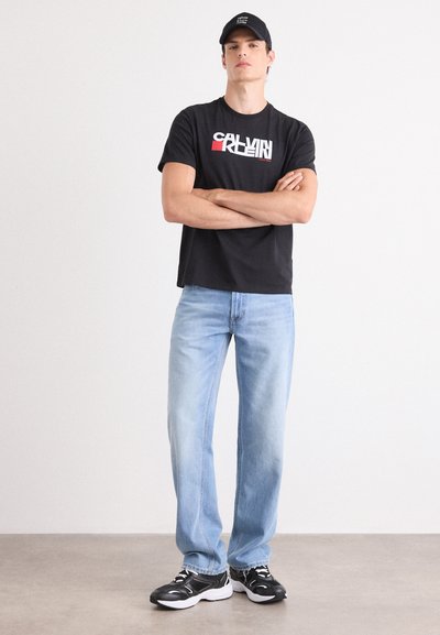 Camiseta negra de algodón con logo de Calvin Klein en blanco y rojo, jeans rectos de mezclilla azul claro y zapatillas negras con acentos en blanco.