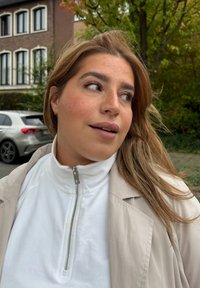 Vit zip-up fleecekrage, beige ytterjacka, vågigt ljusbrunt hår. Mjuk textur, naturligt ljus, grön vegetation och en grå bil i bakgrunden.