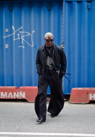 Chaqueta negra, camiseta gráfica, pantalones anchos negros y gafas de sol. De pie cerca de un barrera de concreto rojo y un contenedor de envío azul.
