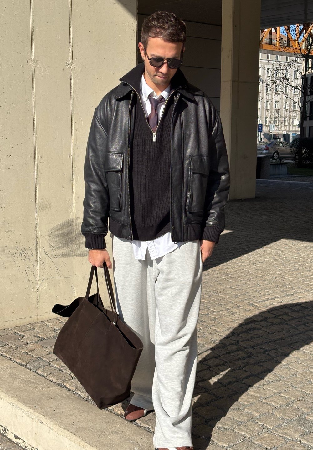 Giubbotto di pelle nera sopra un maglione scuro, pantaloni gris perla e scarpe marroni. Una borsa a tracolla marrone è in mano, con contrasti di luce e ombra visibili.