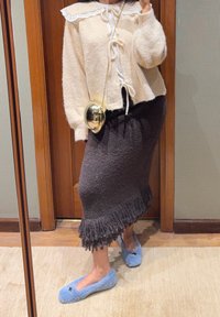 Pull beige clair en tissu duveteux avec bordure en dentelle et ruban à nouer, assorti à une jupe en maille foncée ornée de franges. Chaussures plates bleues duveteuses à enfiler.