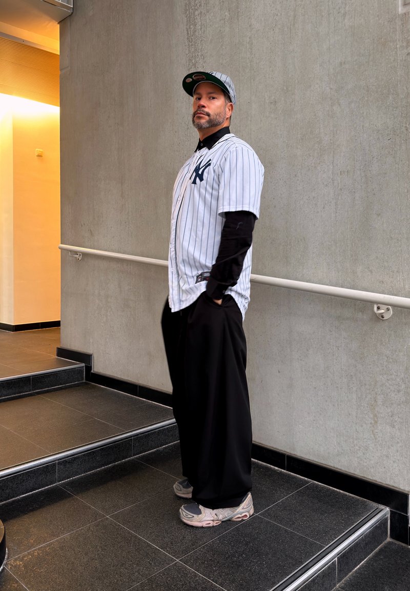 Homme portant un maillot de baseball et une casquette des New York Yankees, un pantalon noir et des baskets, debout sur des escaliers dans un espace intérieur moderne.