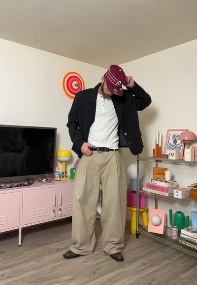 Hombre vistiendo pantalones beige de gran tamaño, chaqueta negra, camisa blanca y gorra burdeos, de pie en una sala de estar moderna y colorida con libros y decoración.