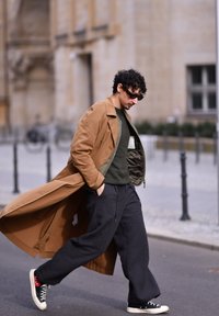 Dlhý hnedý kabát cez olivovo zelený sveter, doplnený tmavými, voľne strihanými nohavicami a čiernymi teniskami. Mesto s rozmazaným pozadím.