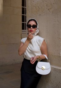 Weiße Lederhandtasche mit goldenen Beschlägen, am Arm getragen. Model in einem cremefarbenen Oberteil, schwarzem Rock und gepunktetem Schal, während sie Lippenstift aufträgt.