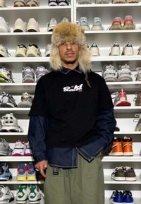 Homme portant un chapeau en fourrure, un t-shirt noir et un pantalon vert se tenant devant des étagères blanches remplies de diverses baskets.