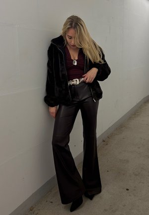 Mujer con cabello rubio largo, vistiendo chaqueta de piel sintética negra, blusa burdeos, cinturón blanco, pantalones de cuero negros de pierna ancha y tacones puntiagudos negros, de pie junto a una pared.