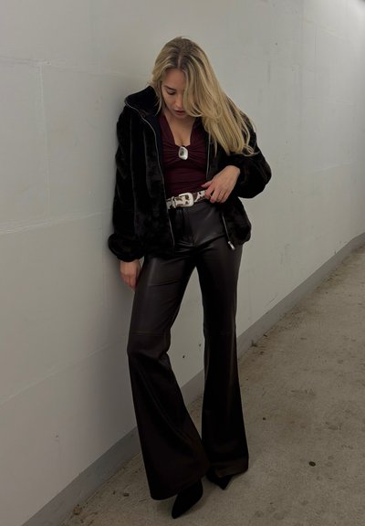 Mujer con cabello rubio largo, vistiendo chaqueta de piel sintética negra, blusa burdeos, cinturón blanco, pantalones de cuero negros de pierna ancha y tacones puntiagudos negros, de pie junto a una pared.