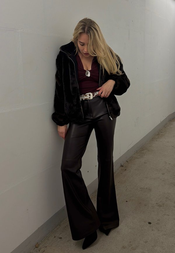 Femme aux longs cheveux blonds portant une veste en fausse fourrure noire, un haut bordeaux, une ceinture blanche, un pantalon large en cuir noir et des talons pointus noirs, debout près d'un mur.