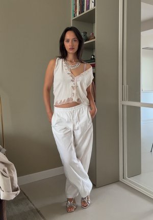 Top blanco corto con detalles de encaje, pantalones blancos holgados y sandalias de tacón plateadas. El fondo presenta una estantería con libros y decoraciones.