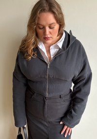 Must kapuutsiga lukuga dressipluus lühendatud lõikega ja soonikkoes varrukaservadega, kombineeritud musta pliiatsseelikuga. Materjal on sile ning kapuutsiga.