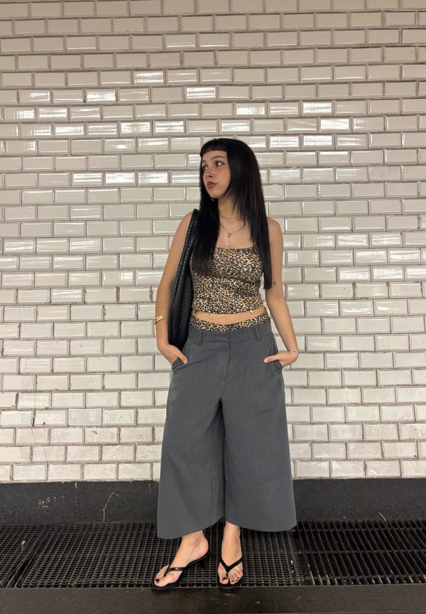 Jeune femme aux longs cheveux foncés, portant un crop top imprimé léopard et un pantalon large gris, debout contre un mur carrelé blanc, les mains dans les poches.