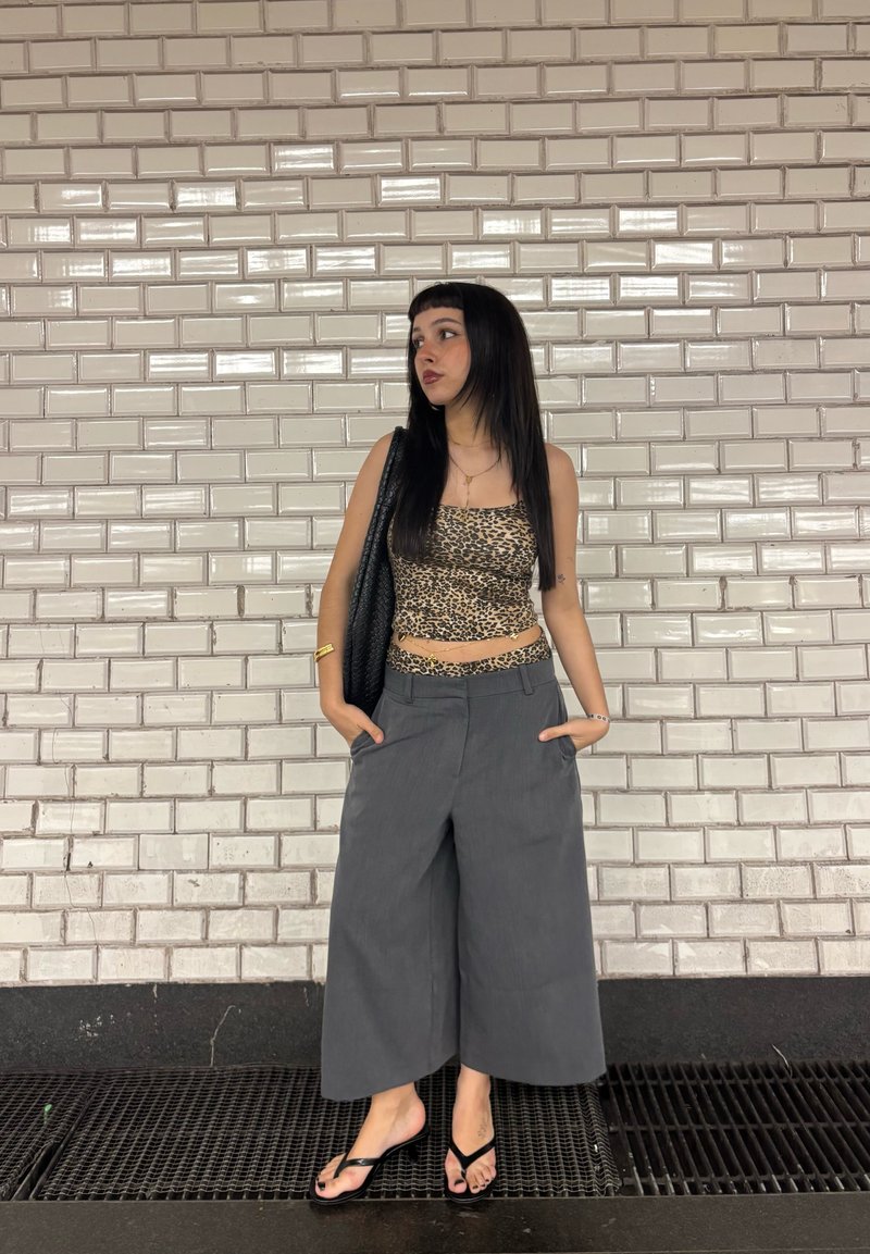 Młoda kobieta o długich ciemnych włosach, ubrana w crop top w panterkę i szerokie szare spodnie, stoi przy białej, kafelkowej ścianie z rękami w kieszeniach.