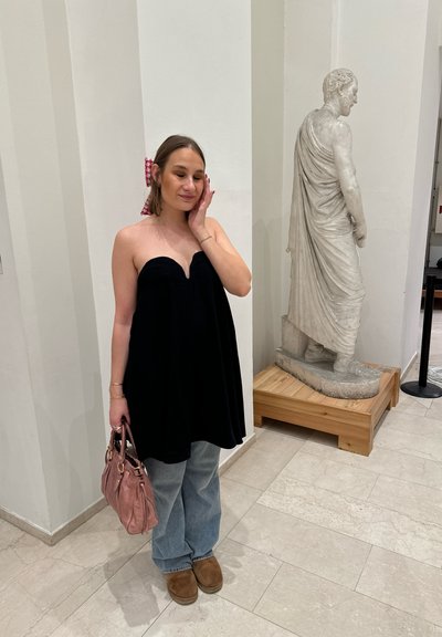 Top negro sin tirantes con forma acampanada, jeans de pierna ancha color azul claro, botas marrones hasta el tobillo y un bolso rosa. Estatua de mármol en el fondo.